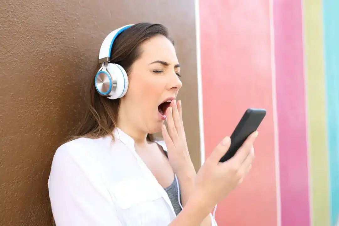 Bored,Woman,Yawning,Using,Smart,Phone,And,Headphones,To,Listen